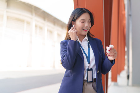 Asian businesswoman enjoying music with wireless earbuds while standing outside a modern office buildingの写真素材