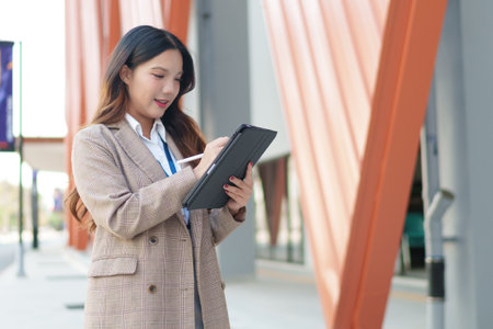 Asian businesswoman working on a tablet while standing outside a modern office buildingの写真素材