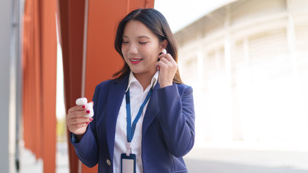 Asian businesswoman enjoying music with wireless earbuds while standing outside a modern office buildingの写真素材