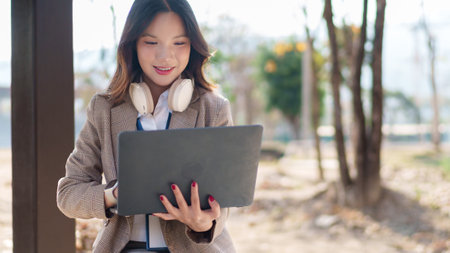 Asian businesswoman working on a laptop while sitting in an outdoor urban setting with headphones around her neckの写真素材