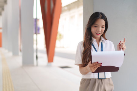 Asian businesswoman multitasking, walking while reading documents outside a modern office buildingの写真素材