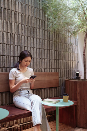 Woman enjoying peaceful coffee break while browsing smartphone at cozy outdoor cafe tableの写真素材