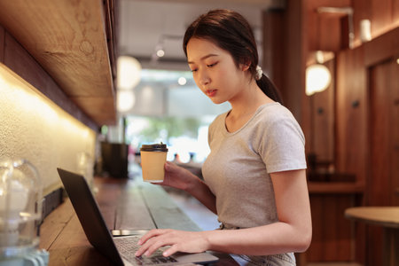Young woman drinking coffee while working on laptop at stylish cafe counter with warm lightingの写真素材