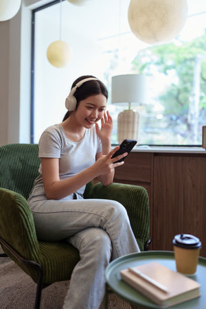Smiling young woman listening to music with wireless headphones while holding smartphone in cafe settingの写真素材