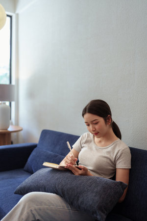 Calm young woman sitting on couch and writing in her diary during a peaceful moment in cozy home settingの写真素材