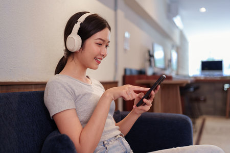 Young woman relaxing on sofa while listening to music with headphones and browsing songs on smartphoneの写真素材