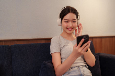 Young woman relaxing on sofa while listening to music with headphones and browsing songs on smartphoneの写真素材