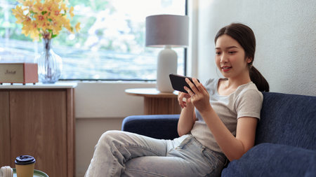 Young woman enjoying mobile gaming on smartphone while sitting on sofa in cozy daylight cafe cornerの写真素材