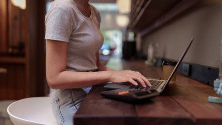 Woman working on laptop and using calculator at cafe counter during casual financial planning sessionの写真素材