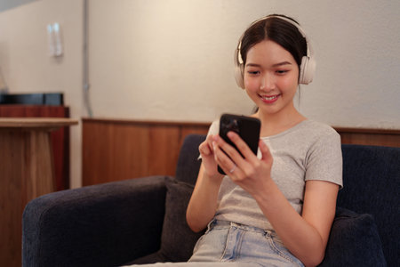 Young woman relaxing on sofa while listening to music with headphones and browsing songs on smartphoneの写真素材
