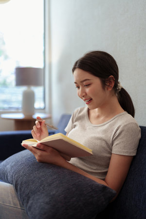 Calm young woman sitting on couch and writing in her diary during a peaceful moment in cozy home settingの写真素材