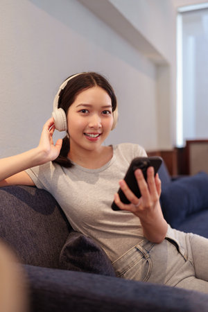 Young woman relaxing on sofa while listening to music with headphones and browsing songs on smartphoneの写真素材