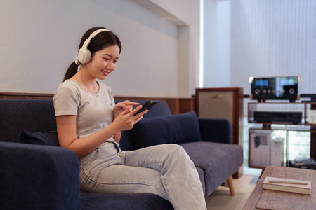 Young woman relaxing on sofa while listening to music with headphones and browsing songs on smartphoneの写真素材
