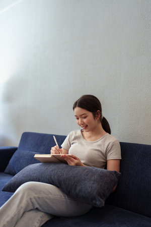 Calm young woman sitting on couch and writing in her diary during a peaceful moment in cozy home settingの写真素材