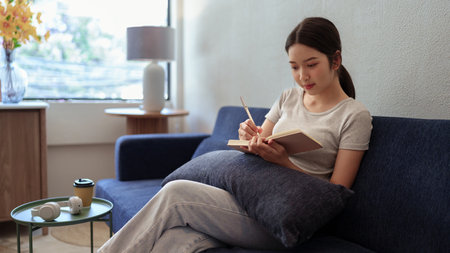 Calm young woman sitting on couch and writing in her diary during a peaceful moment in cozy home settingの写真素材