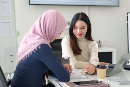 Two young Asian women collaborate on Muslim business plan in a modern office. One wears a hijab while they brainstorm ideas for their startup. Project discussion and strategyの写真素材