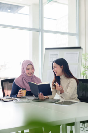 Two young Asian women in a Muslim business discussing a project. They are collaborating on marketing, finance, and startup ideas in a modern office environment.の写真素材