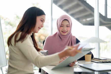Two young Asian women, one wearing a hijab, collaborate on Muslim business startup ideas in a modern office. They analyze data, plan marketing, aiming for business success.の写真素材