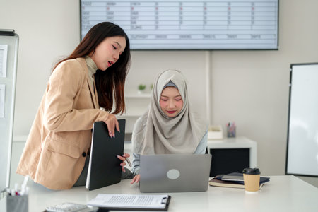 Two young Asian women, one wearing a hijab, collaborate on a laptop in a modern office. Discussing business strategy, financial planning, and teamwork for success.の写真素材