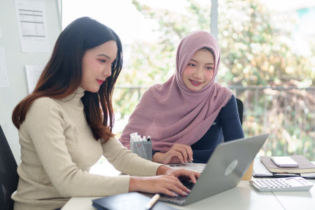 Two young Asian women collaborate on a laptop in a modern office. One woman wears a hijab. They represent Muslim business, teamwork, and entrepreneurship for successful startups.の写真素材