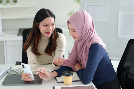 Two adult Asian women collaborate on Muslim business strategies in a bright office. One wears a hijab. They discuss ideas, and entrepreneurship for growth and success in a modern, innovative workspace.の写真素材
