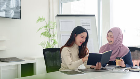 Two young Asian women, one wearing a hijab, collaborate on Muslim business ideas in a modern office setting. Brainstorming and planning for startup success.の写真素材