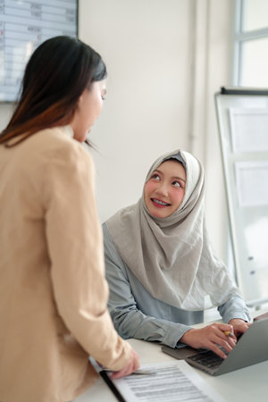 Two young Asian women collaborating in an office. One wearing a hijab, working on a laptop. Teamwork, communication, and modern Muslim professional themes. Ideal for diversity in workplace concepts.の写真素材