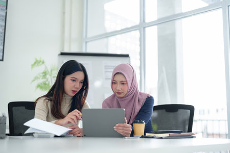 Two young Asian women, one wearing a hijab, collaborate on a tablet at a modern office desk, discussing business ideas and strategy. Muslim business concept.の写真素材