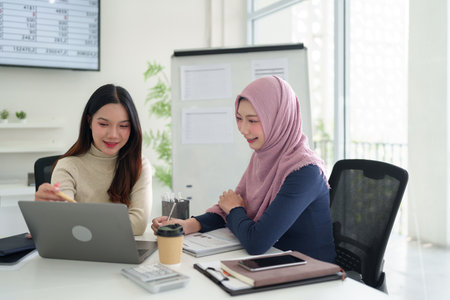 Two young Asian businesswomen, one wearing a hijab, collaborate on a laptop in a bright modern office. Discussing business strategies, analyzing data, and planning for success.の写真素材