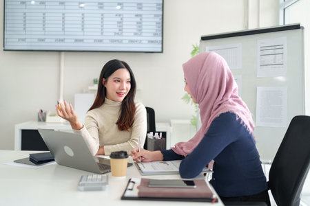 Two young businesswomen, one wearing a hijab, collaborate in a modern office, reviewing charts & discussing Muslim business strategies. Focus on teamwork & Islamic finance growth.の写真素材