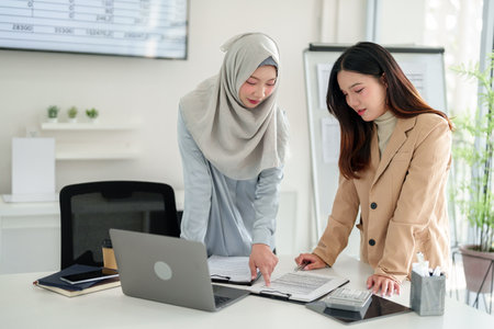 Two young Asian business women collaborate on a contract at an office desk. Reviewing details and strategizing for corporate success with financial data in background.の写真素材
