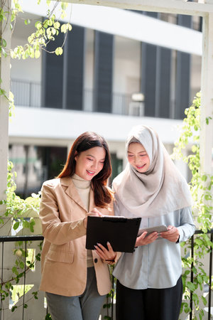 Two young Asian businesswomen are collaborating on a project. One is in a hijab, standing outside in a modern office setting with plants. They are reviewing documents.の写真素材