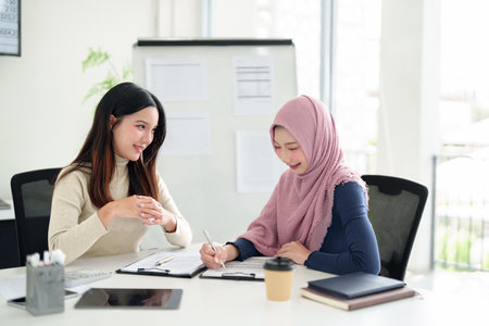 Two young Asian women entrepreneurs, one wearing a hijab, collaborate on Muslim business strategy, reviewing documents in a modern office workspace.の写真素材