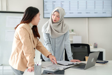 Image of two young Asian women in business attire discussing financial reports. The team collaborates using a laptop, calculator and data for planning. Successful accounting meeting in a modern office.の写真素材