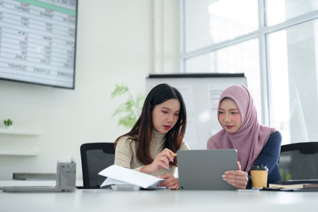 Two young Asian women, one wearing a hijab, collaborate on a Muslim business startup using a digital tablet in a modern office. Highlighting professional teamwork & successの写真素材
