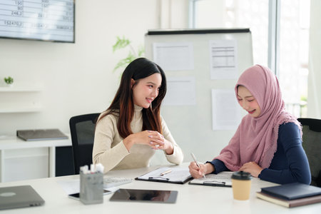 Two Asian women, one wearing a hijab, in a business meeting discussing plans. They represent Muslim business, Islamic finance, and entrepreneurship. This is a collaboration for success.の写真素材