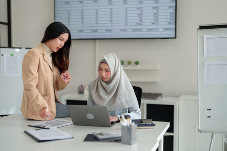 Two young Asian women analyze financial reports in a bright office. One, in a hijab, uses a laptop. Teamwork, collaboration, and professional atmosphere are visible.の写真素材