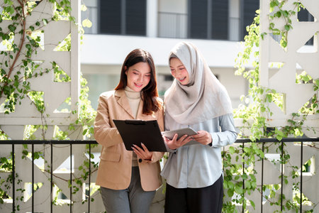 Two young Asian business women are collaborating in a modern office setting, surrounded by lush greenery. They are working together using a tablet and clipboard. Teamwork and discussion.の写真素材