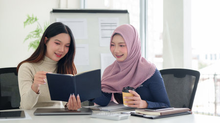 Two young Asian women, one wearing a hijab, collaborate on Muslim business concepts in a bright office. Brainstorming, teamwork, and coffee fuel their startup innovation.の写真素材