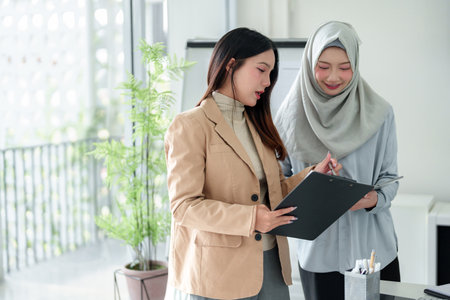 Two Asian women, one wearing a hijab, collaborate on a project in a bright office. They are reviewing documents and discussing business strategies. Image captures teamwork, diversity, and professional success.の写真素材