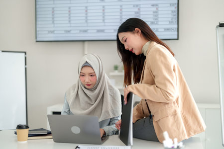 Two adult Asian women in a modern office are collaborating on a laptop. One woman wears a hijab. They are analyzing data charts and discussing work strategy in a professional office setting.の写真素材