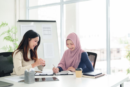 Two young adult Asian women, one wearing a hijab, collaborate on Muslim business strategy financial planning in a bright office. Discussing investment and startup growth.の写真素材