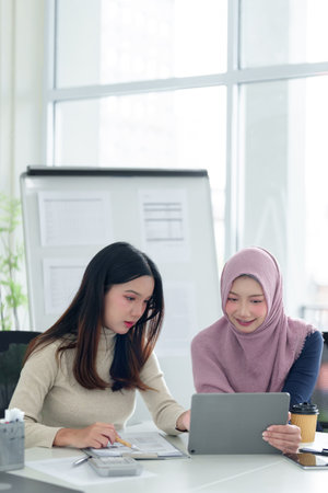 Two young Asian women, one wearing a hijab, collaborate on a Muslim business startup, discussing a project on a tablet in a modern office, showcasing the potential for success.の写真素材