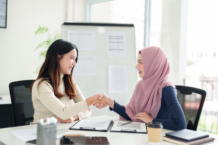 Two young Asian businesswomen, one wearing a hijab, shake hands in a modern office, symbolizing a successful Muslim business partnership, collaboration, and agreement for growth.の写真素材