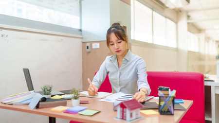 A young Asian woman interior designer is reviewing blueprints at her desk with a laptop, paint...