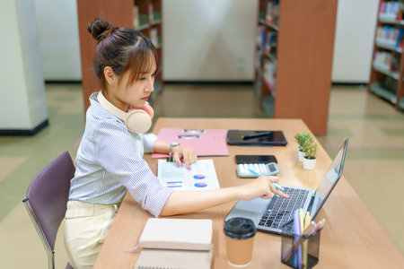Image features a young Asian woman focused on her laptop in a library setting. Ideal for illustrating online education, college life, research, and skill development.の写真素材