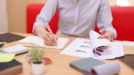 An Asian adult woman interior designer is reviewing blueprints and fabric samples while sketching...