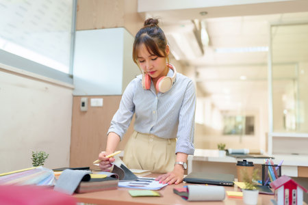 Image of an young Asian woman interior designer reviewing fabric samples and blueprints. Modern office setting.  Focus on design process, creative material selection, home interior projects, and design studio workplace atmosphere.の写真素材