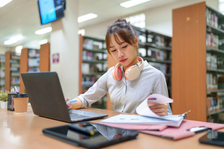 Young Asian woman working on a laptop in a library surrounded by books. She's reviewing documents, researching for a project and furthering her education in college. Focuses on education and learning.の写真素材