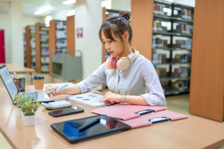 A young Asian woman in a library setting, using a laptop and analyzing charts. Focus on education, research, studying, and data analysis for learning and academic success.の写真素材